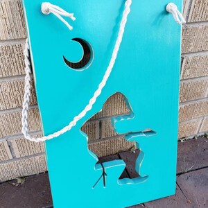 May include: A teal blue wooden sign with a witch silhouette and a crescent moon cutout. The sign has a white rope attached to the top for hanging.