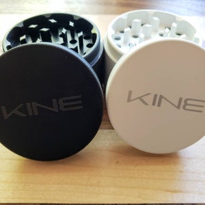 May include: Two metal grinders with black and white lids. Both lids have the word "KINE" printed on them.