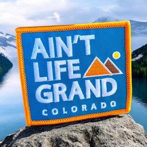 May include: A blue and orange embroidered patch with the text "Ain't Life Grand Colorado" and a graphic of two orange mountains with a yellow sun.