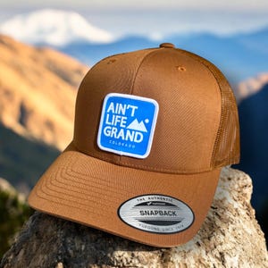 May include: A brown and tan trucker hat with a blue square patch that says "Ain't Life Grand Colorado". The patch has a white outline and a white mountain peak graphic. The hat has a mesh back and a black sticker that says "The Authentic Yupoong Snapback Yupoong Since 1974".