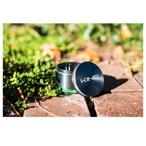 May include: A black metal grinder with a green trim and the word "KINE" on the lid. The grinder is sitting on a brick surface.