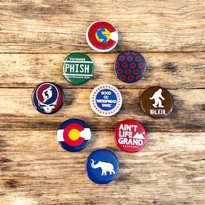 May include: A collection of round pinback buttons with various designs and text. The buttons feature state flags, band logos, and phrases like "WALKIN." and "AIN'T LIFE GRAND COLORADO." The color palette includes red, blue, green, and brown.