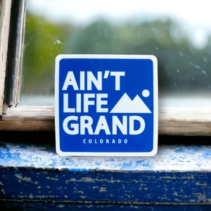 May include: A blue square sticker with white text that reads "Ain't Life Grand Colorado". The sticker features a white mountain silhouette.