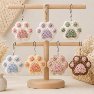 Peut inclure: Sept breloques en forme de patte crochetées suspendues à un présentoir en bois. Chaque breloque présente une base blanche avec un coussinet central coloré et quatre petits coussinets assortis dans des tons pastel ou terre, dont rose, violet, vert, bleu et marron.
