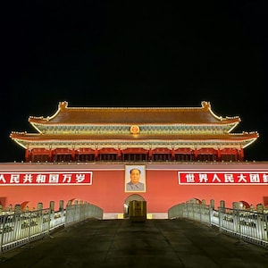 Może przedstawiać: Brama Tiananmen w nocy, z czerwonym murem i centralnym portretem Mao Zedonga. Dwa duże białe banery z czerwonymi chińskimi znakami otaczają portret. Tradycyjny wielopoziomowy dach jest oświetlony żółtymi światłami.