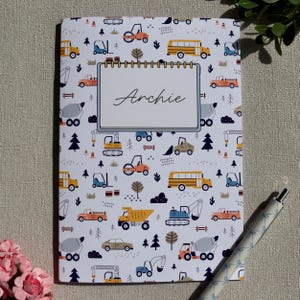 May include: A white notebook featuring a repeating pattern of colorful construction vehicles, including yellow school buses, orange dump trucks, and blue excavators. A central rectangular label with a spiral border displays the name Archie.