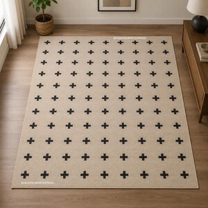 Puede incluir: Una alfombra rectangular con un color base beige claro y un patrón repetitivo de pequeñas cruces negras oscuras dispuestas en cuadrícula. El texto BHADOHICARPETEXPRESS está impreso en letras blancas pequeñas sobre la superficie de la alfombra.