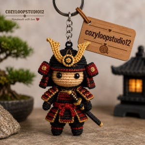 May include: A crocheted samurai keychain featuring a black and red suit of armor with gold accents, a beige face, and a small sword. Attached is a rectangular wooden tag engraved with the text cozyloopstudio12 and a yarn ball icon.