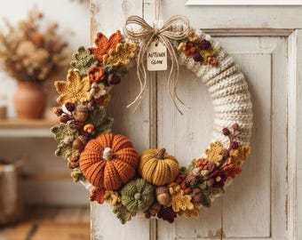 May include: A crochet wreath featuring a cream-colored base adorned with orange, yellow, and green pumpkins, autumn leaves, and small berries. A jute twine bow hangs at the top with a tag labeled "AUTUMN GLOW". The text "DIGITAL PATTERN" is visible.