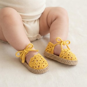 Op de afbeelding: Een paar felgele gehaakte babyschoentjes met een delicaat opengewerkt patroon, een ronde neus en een stevige beige zool in jute-stijl. Elke schoen wordt vastgemaakt met bijpassende gele garen veters die rond de enkel zijn gestrikt.