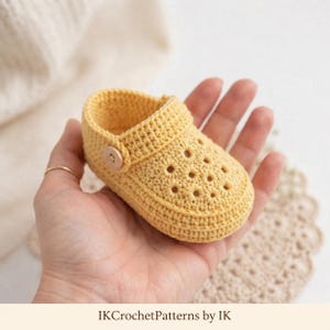 May include: A yellow crochet baby shoe featuring a perforated design on the toe box, a rounded silhouette, and a small wooden button on the side strap. The text IKCrochetPatterns by IK is printed at the bottom of the image.
