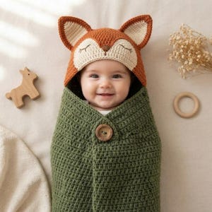May include: A crochet baby swaddle blanket featuring an attached hood designed to look like a fox with orange and cream yarn, pointed ears, and embroidered closed eyes. The swaddle is olive green with a single large wooden button closure at the front.