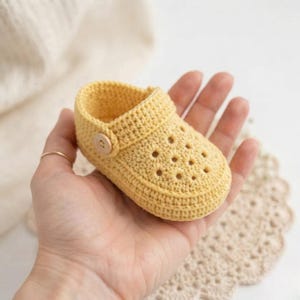 Könnte beinhalten: Ein kleiner, gelber, handgehäkelter Babyschuh mit einem perforierten Muster auf der Oberseite, einer abgerundeten Spitze und einem einzelnen hellen Holzknopf am seitlichen Riemen. Der Schuh ist in einem dichten, strukturierten Maschenbild gefertigt.