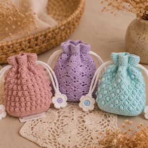 May include: Three small drawstring pouches crocheted in dusty pink, lavender, and light blue yarn. Each bag features a textured pattern and white drawstring cords finished with small white and colored flower-shaped toggles at the ends.