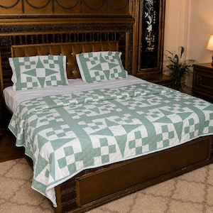 May include: A bed quilt and two matching pillow shams featuring a geometric star and block pattern in sage green and white. The bedding set is laid out flat on a bed, showcasing a repeating quilted design with stitched textures across the fabric.