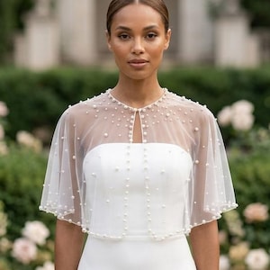May include: A sheer white tulle capelet featuring scattered white pearls throughout the fabric. The capelet has a rounded neckline with a single pearl closure at the center front and falls to the upper arms, worn over a strapless white gown.