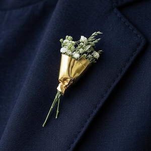 May include: A lapel pin shaped like a miniature bouquet of small white flowers wrapped in a shiny, metallic gold foil cone. The stems of the flowers extend from the bottom of the gold wrapping, and the pin is attached to a dark navy blue blazer.