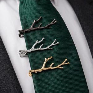 May include: Three tie clips shaped like tree branches in black, silver, and gold tones, attached to a textured dark green necktie. The tie is worn with a crisp white collared shirt and a charcoal gray suit jacket, creating a formal ensemble.