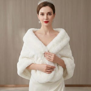 May include: A white faux fur shawl with a thick collar and soft texture, worn over a smooth white garment. The accessory features a wide, draped design that covers the shoulders and upper arms, providing a plush, layered appearance.