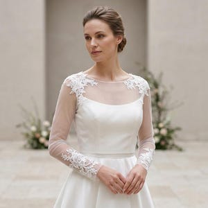 May include: A white floor-length gown featuring a sheer illusion neckline and long sleeves with intricate white floral lace appliqués on the shoulders and cuffs. The bodice is fitted with a smooth fabric, transitioning into a full, flowing skirt.