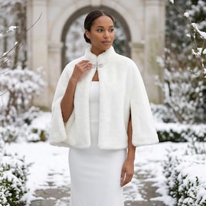 May include: A white faux fur capelet featuring a high collar and a decorative silver-toned clasp at the neckline. The garment is worn over a matching white sleeveless dress, creating a cohesive look with a soft, plush texture and wide sleeves.