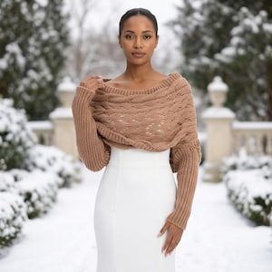 May include: A tan cable-knit shrug featuring a wide, off-the-shoulder neckline and long, ribbed sleeves. The garment is constructed with a textured, chunky knit pattern across the chest and shoulders, paired with a solid white sleeveless dress.