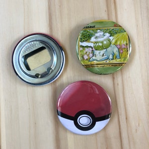 Peut inclure: Trois boutons métalliques ronds. Un bouton est un ouvre-bouteille avec les mots "Bottle Opener" imprimés dessus. Un autre bouton présente une image de dessin animé d'un personnage Pokemon vert avec le texte "Seed Pokemon. Length: 2' 4", Weight: 15 lb" imprimé en dessous. Le troisième bouton est rouge avec un design Pokeball blanc et noir.