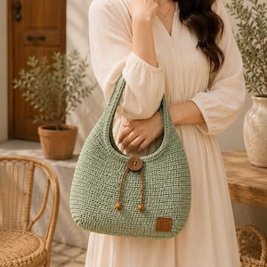 Puede incluir: Un bolso hobo de ganchillo en color verde salvia con un patrón de punto texturizado, una correa de hombro integrada, un cierre central de botón de madera marrón y dos cuentas de madera marrón colgantes. Incluye una pequeña etiqueta cuadrada de cuero sintético marrón.