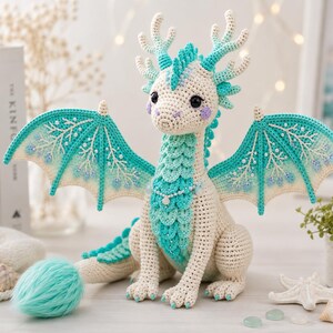 May include: A crocheted dragon toy featuring a cream-colored body with teal accents on the chest, wings, and antlers. The dragon has large black eyes, purple cheek details, and a fluffy teal tail tip, with intricate white embroidery on its wings.