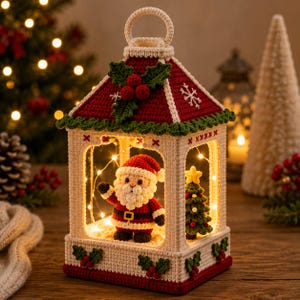 Pode incluir: Uma decoração de lanterna em crochê com um telhado vermelho e branco adornado com folhas de azevinho verdes e bagas vermelhas. Dentro, uma figura de Pai Natal em crochê está ao lado de uma pequena árvore de Natal verde, iluminada por luzes.