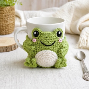 Può includere: Una tazza in ceramica bianca con un rivestimento all'uncinetto verde a forma di rana. La rana ha una pancia bianca rotonda, occhi in plastica nera, guance ricamate in rosa e un sorriso nero. Il rivestimento avvolge la tazza con punti lavorati a maglia e piccole zampe di rana alla base.