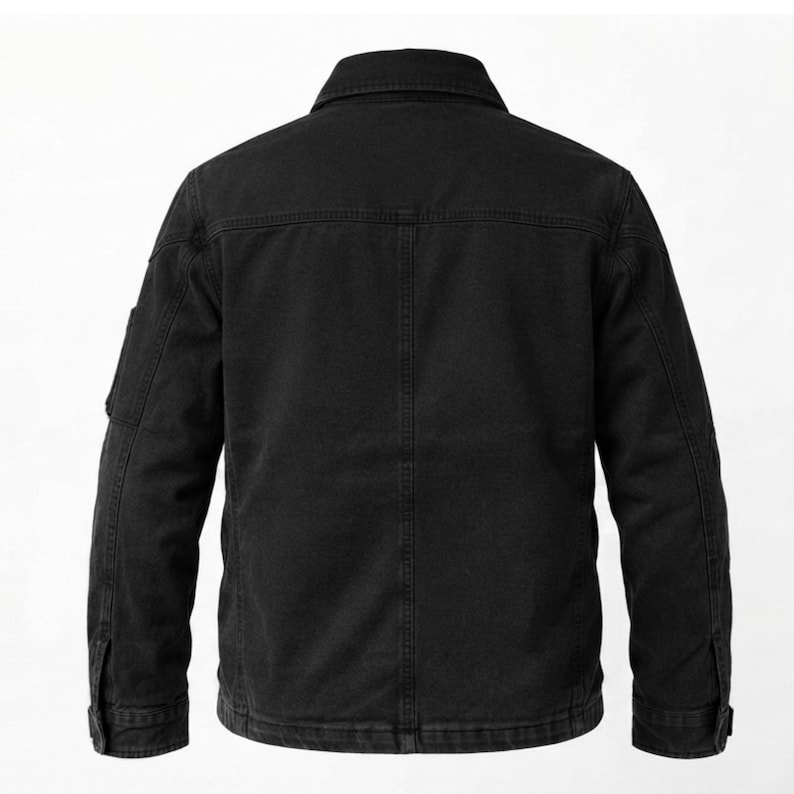 May include: A solid black jacket featuring a structured collar, long sleeves with buttoned cuffs, and a vertical seam running down the center of the back. The garment includes a horizontal yoke across the upper back and a small pocket on the sleeve.