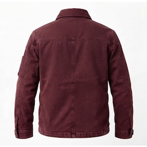 May include: A deep burgundy long-sleeve jacket featuring a structured collar, a horizontal back yoke, and vertical seam detailing. The garment includes long sleeves with buttoned cuffs and a small utility pocket on the upper right sleeve.