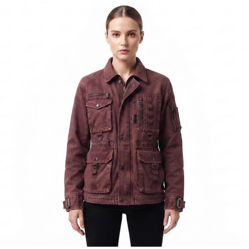 May include: A dark maroon utility jacket featuring a collared neckline, a front button closure, and multiple pockets. The jacket includes two chest pockets, two lower pockets, a zippered sleeve pocket, and metal D-ring hardware accents.