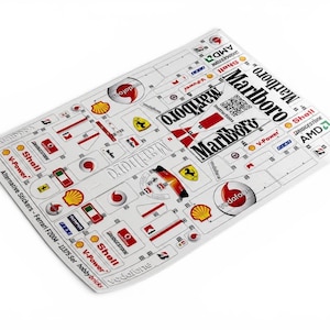 Può includere: Un foglio di adesivi bianchi con vari loghi e decalcomanie per un modello Ferrari F2004. Il foglio include i marchi Marlboro, Vodafone, Shell, Bridgestone e AMD, insieme a emblemi Ferrari, strisce rosse e bianche e un codice QR.