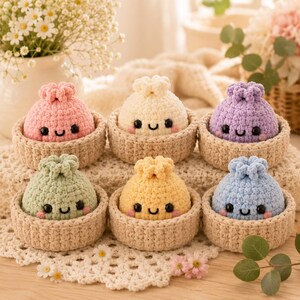 Puede incluir: Seis pequeños juguetes de peluche tejidos a ganchillo, redondos, en colores pastel rosa, crema, morado, verde salvia, amarillo y azul claro. Cada peluche tiene una cara sonriente con ojos negros y mejillas rosadas, dentro de un cuenco beige.