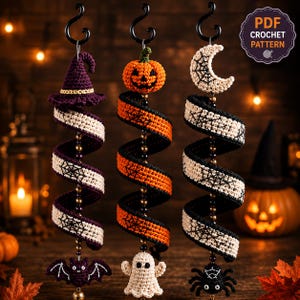 May include: Three hanging crochet spiral decorations featuring Halloween themes. The left has a purple witch hat and bat, the centre an orange pumpkin and ghost, and the right a white crescent moon and spider. Each hangs from a black hook.