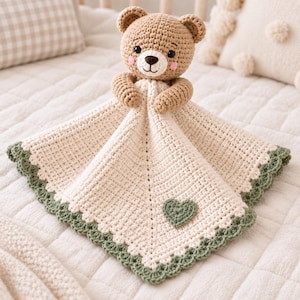 Pode incluir: Uma manta de segurança de crochê com uma cabeça de urso castanho-claro, olhos pretos e bochechas cor-de-rosa. A manta anexada é de cor creme com um ponto texturizado, um pequeno coração verde aplicado no centro e uma borda festonada verde.