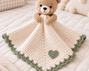 May include: A crochet security blanket featuring a light brown bear head with black eyes and pink cheeks. The attached blanket is cream-colored with a textured stitch, a small green heart appliqué in the center, and a scalloped green border edge.