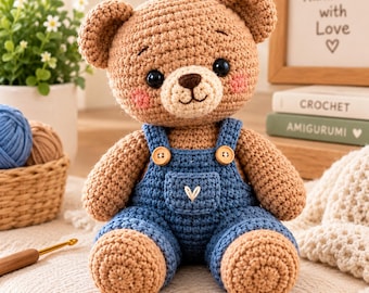 May include: A crocheted teddy bear with light brown yarn, wearing blue overalls with two wooden buttons and a small white embroidered heart on the front pocket. The bear has black safety eyes, a dark brown stitched nose, and pink blush cheeks.