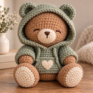 Könnte beinhalten: Ein gehäkelter brauner Teddybär, der einen salbeigrünen Kapuzenpullover mit einem cremefarbenen Herzaufnäher auf der Vordertasche trägt. Der Bär hat eine cremefarbene Schnauze, dunkelbraun gestickte Augen und passende cremefarbene Flecken an den Füßen.