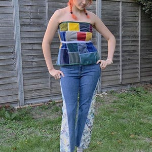 Puede incluir: Un top palabra de honor de patchwork con cuadrados de tela amarilla, roja, azul y gris con una banda horizontal blanca, combinado con vaqueros azules que tienen paneles laterales anchos decorados con un estampado floral claro y zapatos negros.