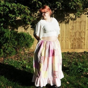 Op de afbeelding: Een wit kort topje met korte mouwen, een ruches bij de halslijn en een gesmokte taille, gecombineerd met een lange patchwork rok tot op de enkels. De rok heeft een herhalend patroon van lichtroze, gele en bloemrijke stoffen vierkanten.