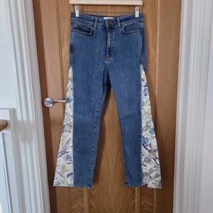 May include: Blue denim jeans featuring a high-waisted design with a button and zipper fly closure. The legs are accented with wide, triangular side panels made of white fabric decorated with a multi-colored floral pattern in blue, green, and yellow.