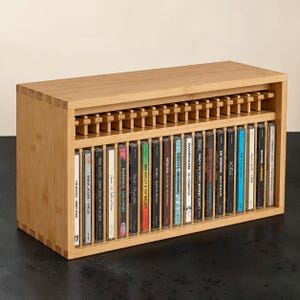 May include: A rectangular bamboo storage rack with finger-jointed corners, designed to hold a single row of compact discs. The rack features a series of vertical dividers and a top shelf, currently filled with twenty-one assorted music albums.