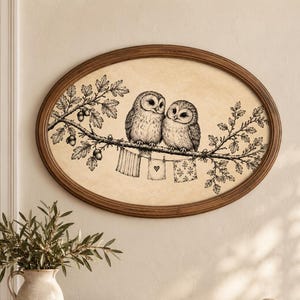 May include: An oval-framed wall art piece featuring a black and white ink illustration of two owls perched on an oak branch with acorns. Below the branch, three small pieces of fabric hang from a clothesline, including one with a heart design.