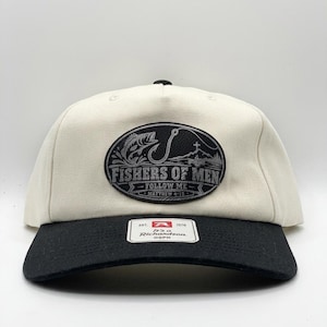May include: A cream-colored baseball cap with a black brim and a black button on top. An oval patch on the front features a fish, a fishing hook, a cross, and the text "FISHERS OF MEN - FOLLOW ME - MATTHEW 4:19". A white tag on the brim reads "Richardson".
