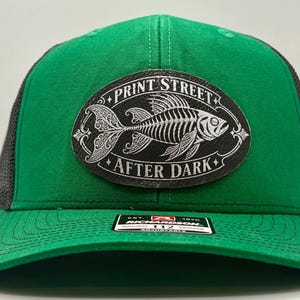 May include: A green and black trucker hat featuring a black oval patch on the front. The patch displays a white fish skeleton illustration with the text PRINT STREET and AFTER DARK. A black sticker is on the brim with Richardson 112 branding.