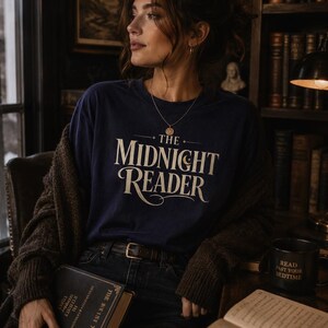 May include: A navy blue long-sleeve t-shirt featuring the cream-coloured text "THE MIDNIGHT READER" printed on the chest. The shirt is worn with a dark brown knit cardigan, a black belt, and dark jeans, while holding a hardback book.