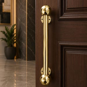 May include: A polished gold-tone metal door handle featuring a long cylindrical bar with spherical ends. The handle is mounted vertically onto a dark brown wooden door with recessed paneling, showcasing a reflective and smooth metallic finish.